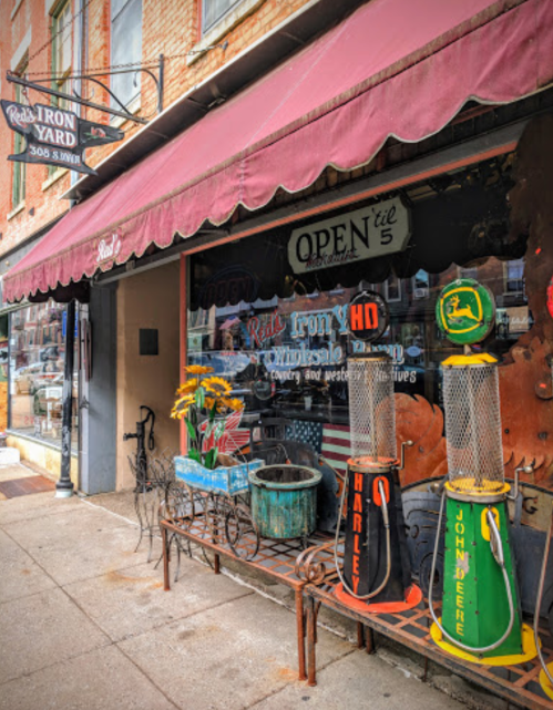 A vintage storefront with a maroon awning, featuring old gas pumps and flower pots outside.