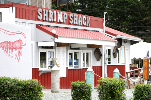 A red and white building with a sign reading "Shrimp Shack," featuring outdoor seating and a mural of a shrimp.