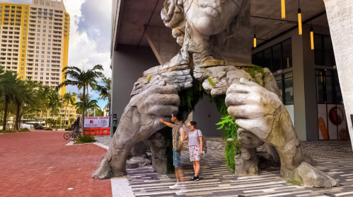 Two people stand in front of a large, cracked sculpture of a figure emerging from a building, surrounded by greenery.