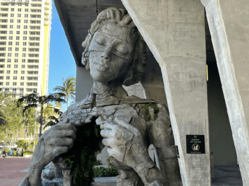 A large, abstract sculpture of a woman with a serene expression, partially covered in greenery, under a concrete structure.