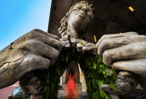 A large, abstract sculpture of a figure breaking through a green, moss-covered frame, set against a modern building.