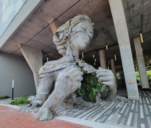 A large, abstract sculpture of a woman holding greenery, set against a modern architectural backdrop.