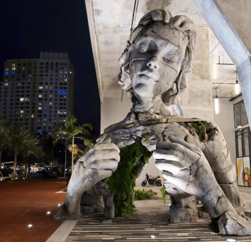 A large, abstract sculpture of a woman with a cracked surface, surrounded by greenery, under a night sky.