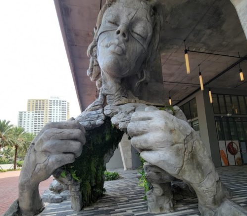 A large, abstract sculpture of a woman with a hollow chest, surrounded by greenery and modern architecture.