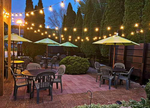 Outdoor dining area with tables, green umbrellas, and string lights, surrounded by tall hedges at dusk.