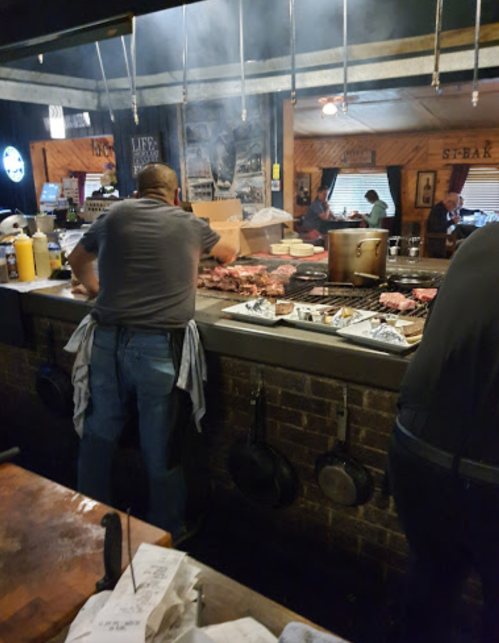 A chef prepares food at a busy grill station, with smoke and various dishes visible in a rustic restaurant setting.