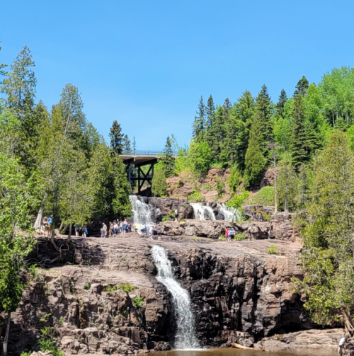 A scenic view of waterfalls cascading over rocky terrain, surrounded by lush green trees and a clear blue sky.