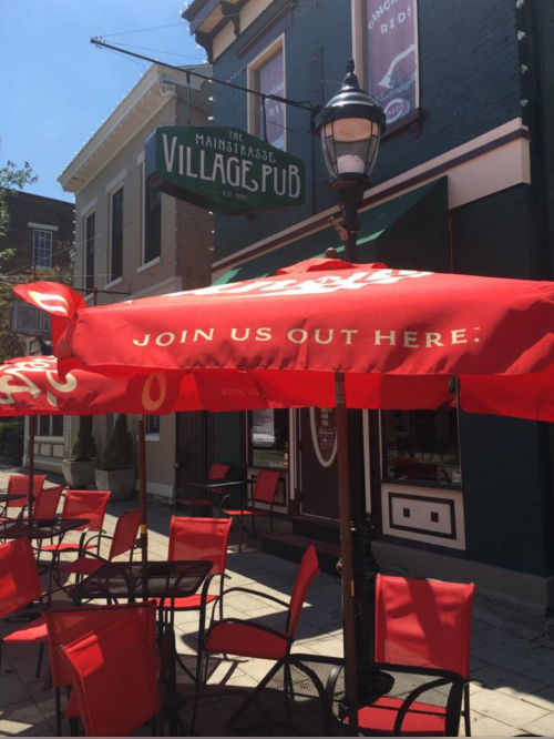 A red umbrella with "Join Us Out Here" text shades outdoor seating at Village Pub, a charming street-side restaurant.