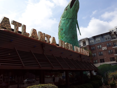 A large green fish sculpture towers over the Atlanta Fish Market restaurant, with a blue sky in the background.