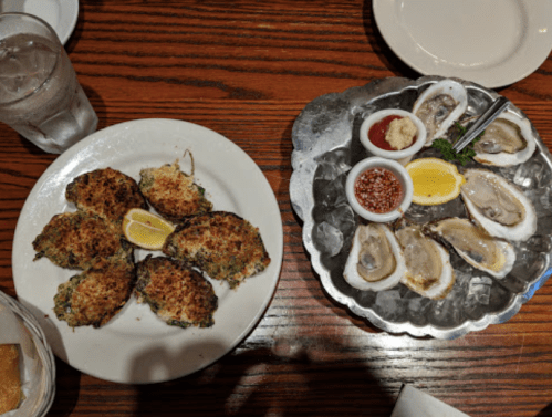A plate of baked oysters with lemon and a platter of raw oysters with sauces on a wooden table.