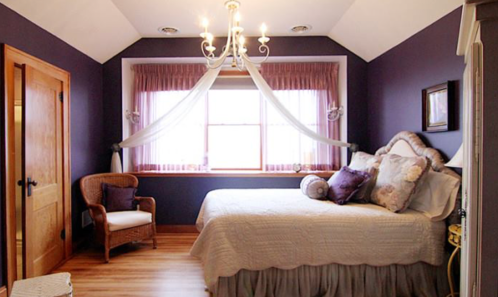 Cozy bedroom with a purple accent wall, a bed with decorative pillows, and a chandelier above a window with sheer curtains.