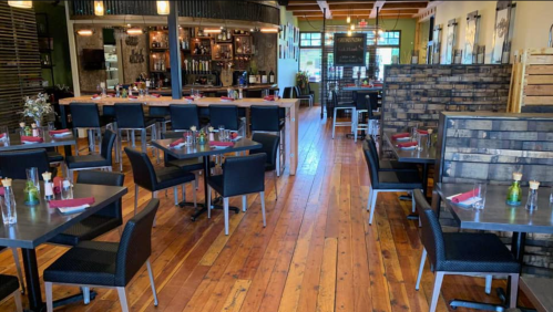 Interior of a modern restaurant featuring wooden floors, tables set for dining, and a bar area in the background.