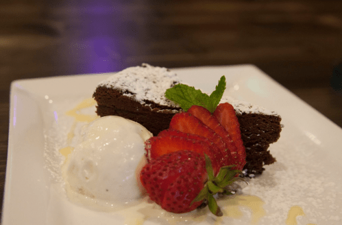 A slice of chocolate cake dusted with powdered sugar, served with a scoop of vanilla ice cream and fresh strawberries.