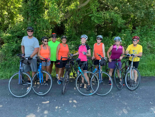 A group of eight cyclists in bright clothing poses with their bikes on a sunny day, surrounded by greenery.