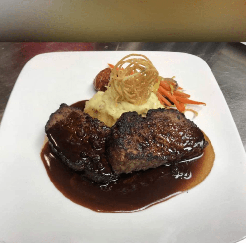 A plate of grilled meat with a rich sauce, served with mashed potatoes, carrots, and crispy onion strings on top.