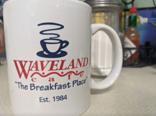 A white coffee mug with the logo of Waveland Cafe, reading "The Breakfast Place" and established in 1984.