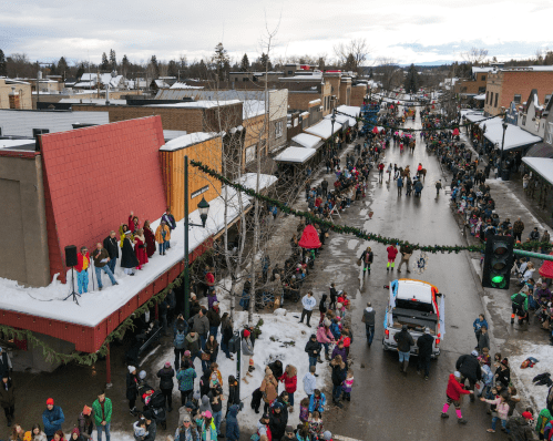 A festive street scene with a crowd gathered for a parade, featuring performers on a stage and snow-covered buildings.