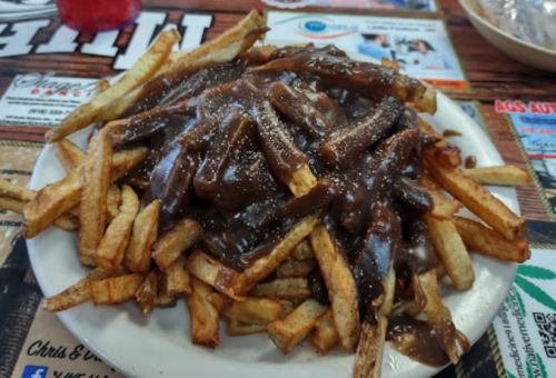 A plate of fries topped with a rich brown gravy, served on a wooden table with various advertisements in the background.