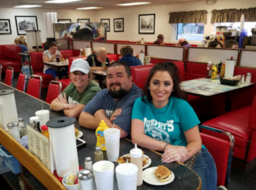 Three people sit at a diner counter, smiling, with food and drinks in front of them. Red booths and patrons in the background.