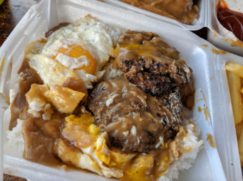 A styrofoam container with rice topped with a hamburger patty, gravy, fried egg, and pieces of chicken.