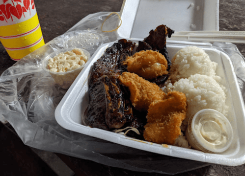 A takeout meal featuring grilled meat, fried chicken, rice, macaroni salad, and a drink in a plastic cup.