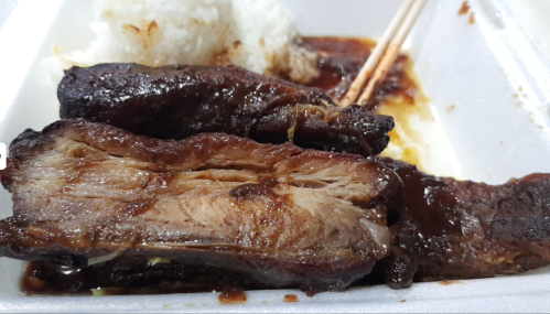 Close-up of tender, glazed beef ribs with rice in a takeout container, chopsticks resting on the side.