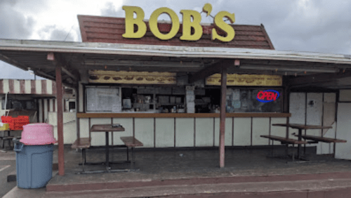 A small food stand named "Bob's" with an open sign, featuring outdoor seating and a pink trash bin nearby.
