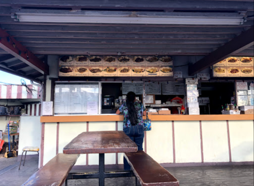 A person stands at a food counter with a menu displayed above, in a casual outdoor dining setting.