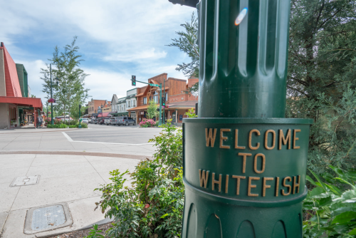 A green street sign reads "Welcome to Whitefish" with a view of a charming downtown street in the background.