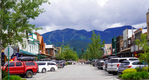 A scenic street view of a small town with shops, parked cars, and mountains in the background under a cloudy sky.