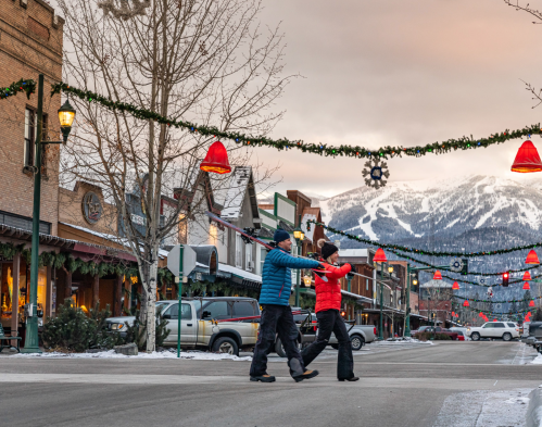 Two people walk hand in hand down a festive street decorated with holiday lights and snow-capped mountains in the background.