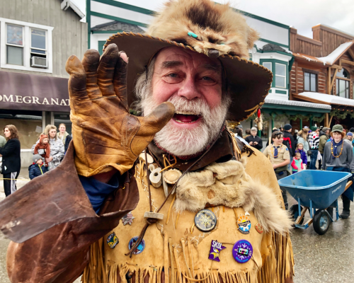 A smiling man in a fur hat and leather gloves waves, adorned with badges, at a festive outdoor event.
