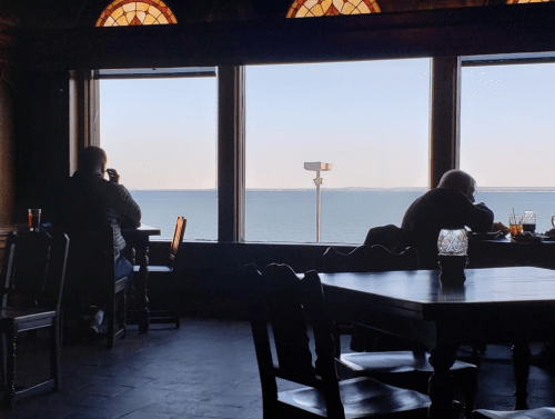 Two people sit at a table in a restaurant, gazing out at a calm sea through large windows.