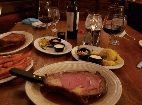 A plate of prime rib with sides of sweet potato fries, vegetables, and sauces, accompanied by glasses of red wine.