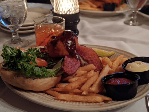 A plate with a burger topped with barbecue sauce, lettuce, and tomato, served with fries and dipping sauces.