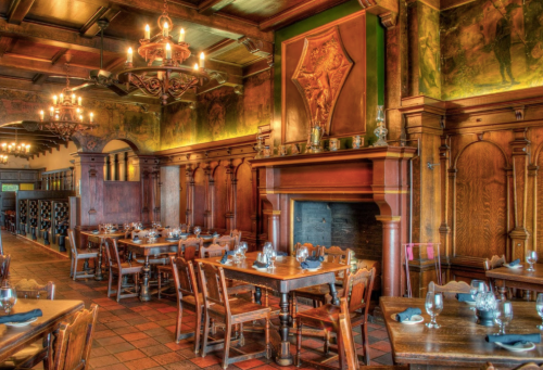 Cozy restaurant interior with wooden tables, chairs, and ornate decor, featuring a large fireplace and chandeliers.