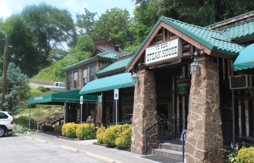 Exterior of a rustic steakhouse with green awnings, stone walls, and landscaped parking area.