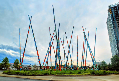 Colorful, tall poles arranged in a circular pattern, surrounded by greenery and a cloudy sky in the background.