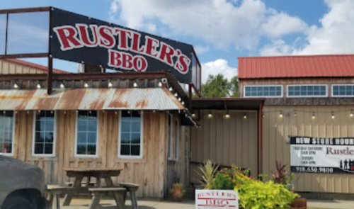 Exterior of Rustler's BBQ restaurant with a wooden facade, sign, and outdoor seating under a blue sky.