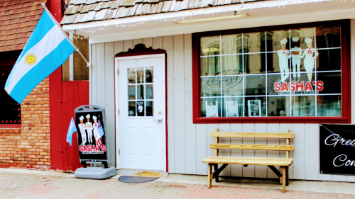 Exterior of Sasha's Empanadas with an Argentine flag, open sign, and a bench in front.