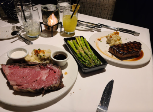 A table setting with two plates: one with prime rib, mashed potatoes, and sauce; the other with grilled steak and asparagus.