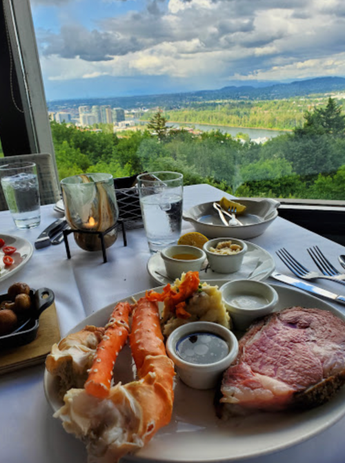 A gourmet meal featuring prime rib, lobster, and sides, with a scenic view of a city and mountains in the background.