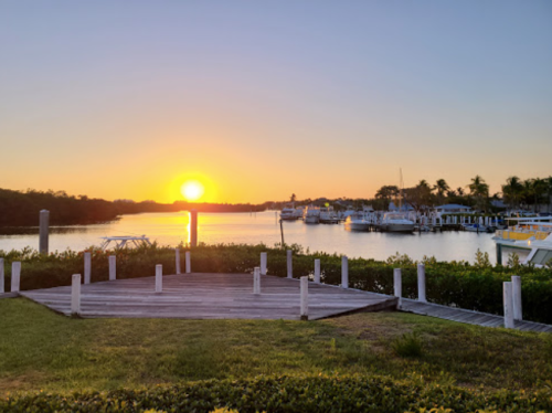 Sunset over a calm waterway, with boats docked and lush greenery in the foreground.