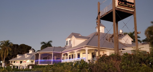 A waterfront restaurant with a white exterior and a metal roof, surrounded by palm trees at dusk.