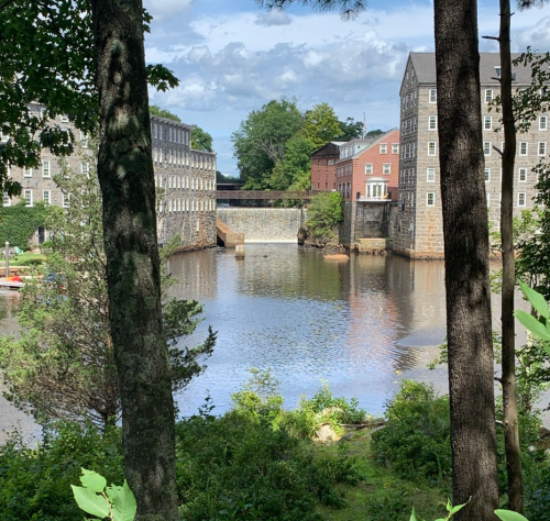 A serene river scene framed by trees, featuring historic buildings and a dam under a partly cloudy sky.