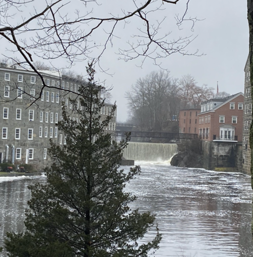 A river flows past historic buildings and a waterfall, surrounded by bare trees on a cloudy day.