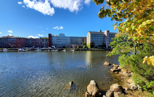 A serene river scene with buildings along the shore and colorful autumn leaves in the foreground under a clear blue sky.