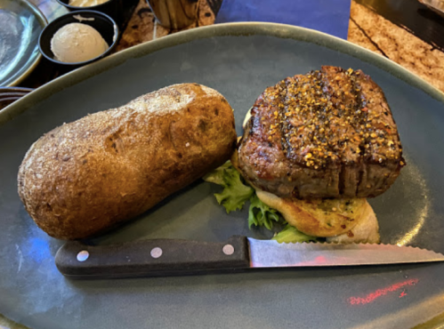 A plate with a grilled steak, seasoned and resting on lettuce, alongside a baked potato and a knife.