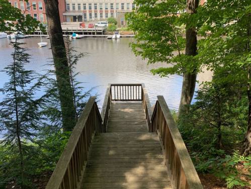 A wooden walkway leads to a calm river, surrounded by trees and buildings in the background.