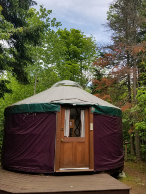 A cozy yurt with a wooden door, surrounded by lush green trees under a partly cloudy sky.